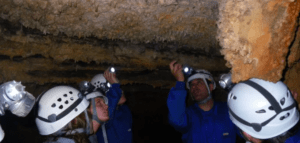 Aventureros con casco exploran una cueva de Montanejos, iluminando estalactitas con linternas mientras participan en "Espeleología con Rápel en Montanejos". - ForTwo - Planes para parejas