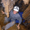 Una persona con equipo completo y casco sonríe de emoción mientras se agacha en un estrecho y rocoso pasaje de una cueva, lista para explorar las maravillas de la Espeleología con Rápel en Montanejos. - ForTwo - Planes para parejas