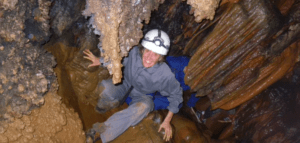 Una persona con equipo completo y casco sonríe de emoción mientras se agacha en un estrecho y rocoso pasaje de una cueva, lista para explorar las maravillas de la Espeleología con Rápel en Montanejos. - ForTwo - Planes para parejas