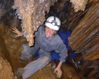 Una persona con equipo completo y casco sonríe de emoción mientras se agacha en un estrecho y rocoso pasaje de una cueva, lista para explorar las maravillas de la Espeleología con Rápel en Montanejos. - ForTwo - Planes para parejas