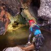 Vestido con equipo de espeleología, una persona explora un pasaje de cueva rocoso lleno de agua con una linterna frontal, abrazando la aventura de "Espeleología en Valencia: una aventura subterránea para vivir en pareja". - ForTwo - Planes para parejas