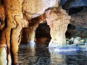 La entrada de la cueva iluminada por el sol con paredes rocosas se refleja en el agua tranquila, conduciendo a un interior más oscuro, perfecto para experimentar "Espeleología en Valencia: una aventura subterránea para vivir en pareja". - ForTwo - Planes para parejas