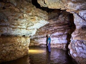 Con casco puesto, alguien se embarca en "Espeleología en Valencia: una aventura subterránea para vivir en pareja", explorando una cueva rocosa. El agua brilla bajo los pies, reflejando el haz de luz de su linterna mientras se adentran en este cautivador mundo subterráneo. - ForTwo - Planes para parejas