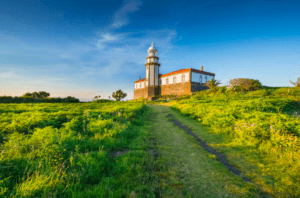 Bajo un cielo azul al atardecer, el faro es un faro para aquellos que realizan la Excursión en Barco a la Isla de Ons, ofreciendo impresionantes vistas al mar y al horizonte desde su posición elevada en la colina cubierta de hierba. - ForTwo - Planes para parejas