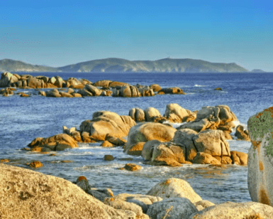 Costas rocosas con un mar azul, grandes rocas dispersas y colinas bajas distantes bajo un cielo despejado lo hacen perfecto para una "Excursión en Barco a la Isla de Ons". - ForTwo - Planes para parejas