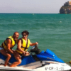 Dos personas en una moto acuática "Explora el Mediterráneo en moto de agua", ataviadas con chalecos salvavidas, se deslizan por la bahía con un impresionante castillo costero al fondo, capturando la esencia de la aventura mediterránea. - ForTwo - Planes para parejas