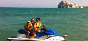 Dos personas en una moto acuática "Explora el Mediterráneo en moto de agua", ataviadas con chalecos salvavidas, se deslizan por la bahía con un impresionante castillo costero al fondo, capturando la esencia de la aventura mediterránea. - ForTwo - Planes para parejas