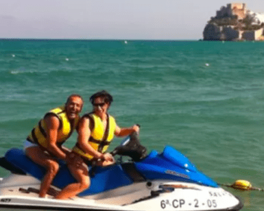 Dos personas en una moto acuática "Explora el Mediterráneo en moto de agua", ataviadas con chalecos salvavidas, se deslizan por la bahía con un impresionante castillo costero al fondo, capturando la esencia de la aventura mediterránea. - ForTwo - Planes para parejas