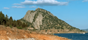 Bajo un cielo parcialmente nublado, la colina rocosa con escasa vegetación contempla las serenas aguas azules del Embalse de Alange. Es un paisaje que invita a explorar, ya sea haciendo senderismo o embarcándose en la aventura "Explora el Embalse de Alange en piragua". - ForTwo - Planes para parejas