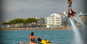 Una persona utiliza con destreza el Flyboard en Alcocéber sobre aguas cristalinas cerca de una moto acuática, con los encantadores edificios de Alcocéber y las playas de arena creando un fondo pintoresco. - ForTwo - Planes para parejas
