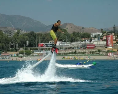 Alguien está disfrutando del Flyboard en Marbella, volando sobre el océano con el impresionante telón de fondo de las montañas y los edificios de Marbella que realzan la impresionante escena. - ForTwo - Planes para parejas