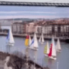 Los veleros se deslizan elegantemente por el río bajo un majestuoso puente colgante, con los encantadores edificios de Bilbao alineando las orillas, ofreciendo una experiencia inolvidable con "Iniciación a la vela en Bilbao. - ForTwo - Planes para parejas