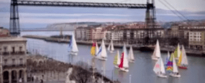 Los veleros se deslizan elegantemente por el río bajo un majestuoso puente colgante, con los encantadores edificios de Bilbao alineando las orillas, ofreciendo una experiencia inolvidable con "Iniciación a la vela en Bilbao. - ForTwo - Planes para parejas