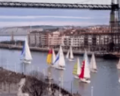 Los veleros se deslizan elegantemente por el río bajo un majestuoso puente colgante, con los encantadores edificios de Bilbao alineando las orillas, ofreciendo una experiencia inolvidable con "Iniciación a la vela en Bilbao. - ForTwo - Planes para parejas