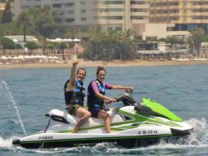 Dos personas viajan en una moto acuática en Marbella, una de ellas haciendo un gesto de aprobación con el pulgar hacia arriba, frente al horizonte de la ciudad. Disfrutando de emocionantes deportes acuáticos, sonríen entre las olas iluminadas por el sol. - ForTwo - Planes para parejas