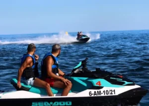 En Marbella, dos personas en una "Moto de agua en Marbella" observan a otra persona cruzar el agua a toda velocidad en la distancia bajo un cielo despejado. - ForTwo - Planes para parejas