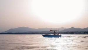 Un pequeño barco navega por aguas tranquilas al atardecer, con vistas a las majestuosas montañas. Experimente esta serena escena con "Navega sin Licencia en Marbella", donde podrá navegar sin licencia en estas tranquilas aguas. - ForTwo - Planes para parejas