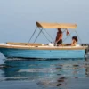 Una pequeña lancha motora azul con toldo beige se desliza por las tranquilas aguas de Marbella, transportando a un hombre de pie y a una mujer sentada disfrutando de la libertad que ofrece "Navega sin Licencia en Marbella". - ForTwo - Planes para parejas