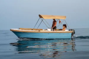Una pequeña lancha motora azul con toldo beige se desliza por las tranquilas aguas de Marbella, transportando a un hombre de pie y a una mujer sentada disfrutando de la libertad que ofrece "Navega sin Licencia en Marbella". - ForTwo - Planes para parejas