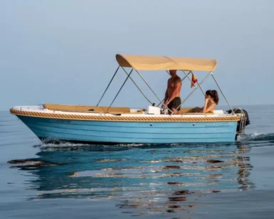 Una pequeña lancha motora azul con toldo beige se desliza por las tranquilas aguas de Marbella, transportando a un hombre de pie y a una mujer sentada disfrutando de la libertad que ofrece "Navega sin Licencia en Marbella". - ForTwo - Planes para parejas