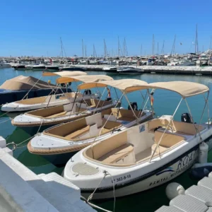 Cuatro lanchas motoras vacías con toldos color canela están atracadas en un puerto deportivo de Marbella, rodeadas de veleros. Experimenta la libertad de explorar estas aguas sin esfuerzo con "Navega sin Licencia en Marbella" y haz realidad tus sueños náuticos sin necesidad de licencia. - ForTwo - Planes para parejas