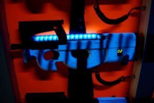 Descubre la emocionante Oferta: 2 partidas de Laser Tag en Alfafar con una pistola futurista con luces azules y pantalla digital, todo sobre un fondo naranja vibrante, ¡perfecto para juegos llenos de adrenalina!. - ForTwo - Planes para parejas