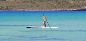 Una persona se arrodilla sobre un Paddle Surf en el Embalse del Sichar, deslizándose por aguas azules claras con una exuberante costa verde que crea un pintoresco telón de fondo. - ForTwo - Planes para parejas