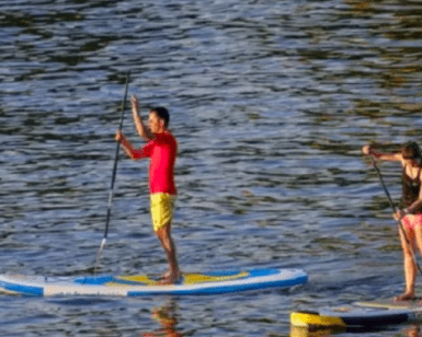 En el Embalse del Sichar, dos personas practican paddle surf, uno viste camiseta roja y pantalón corto amarillo, mientras que el otro viste ropa oscura. - ForTwo - Planes para parejas