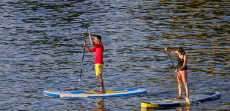 En el Embalse del Sichar, dos personas practican paddle surf, uno viste camiseta roja y pantalón corto amarillo, mientras que el otro viste ropa oscura. - ForTwo - Planes para parejas