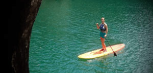 Utilizando el Paddle Surf en el Embalse del Sichar, un paddleboarder se desliza sobre serenas y claras aguas verdes junto a una cornisa rocosa, abrazando una pacífica aventura. - ForTwo - Planes para parejas