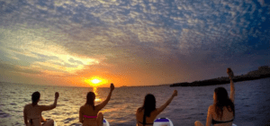 Cuatro personas en la experiencia "Paddle surf romántico en Menorca" disfrutan de una puesta de sol sobre el océano, cada una levantando un brazo hacia el cielo al unísono. - ForTwo - Planes para parejas