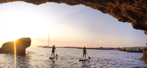El Paddle surf romántico en Menorca presenta a personas deslizándose cerca de la entrada de una cueva al atardecer, con un velero a lo lejos realzando la belleza serena y romántica de Menorca. - ForTwo - Planes para parejas