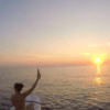 Dos personas en un barco saludan en una romántica puesta de sol sobre el tranquilo océano cerca de Menorca, con "Paddle surf romántico en Menorca" mostrando un velero deslizándose con gracia en la distancia. - ForTwo - Planes para parejas