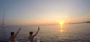 Dos personas en un barco saludan en una romántica puesta de sol sobre el tranquilo océano cerca de Menorca, con "Paddle surf romántico en Menorca" mostrando un velero deslizándose con gracia en la distancia. - ForTwo - Planes para parejas