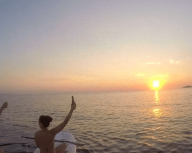 Dos personas en un barco saludan en una romántica puesta de sol sobre el tranquilo océano cerca de Menorca, con "Paddle surf romántico en Menorca" mostrando un velero deslizándose con gracia en la distancia. - ForTwo - Planes para parejas