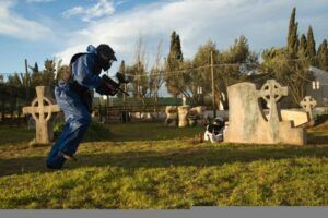 Vestido con uniforme azul, alguien se lanza a la batalla definitiva de paintball, sorteando estructuras de piedra y árboles en los pintorescos campos de Valencia bajo un cielo parcialmente nublado. ¡Vívelo con "Paintball en Valencia: ¡vive la batalla definitiva!. - ForTwo - Planes para parejas