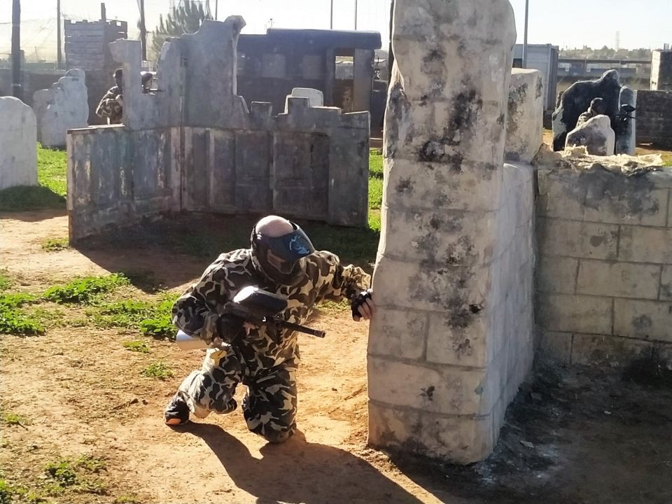 En el corazón de Valencia, alguien camuflado se agacha con una pistola de paintball tras una barrera de hormigón, inmerso en "Paintball en Valencia: ¡vive la batalla definitiva!" en un campo al aire libre. - ForTwo - Planes para parejas