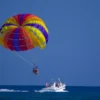Una persona disfruta de la experiencia de Parasailing en Marbella, volando con un paracaídas vibrante, remolcado por un barco blanco sobre el océano brillante bajo un cielo azul claro. - ForTwo - Planes para parejas