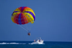 Una persona disfruta de la experiencia de Parasailing en Marbella, volando con un paracaídas vibrante, remolcado por un barco blanco sobre el océano brillante bajo un cielo azul claro. - ForTwo - Planes para parejas