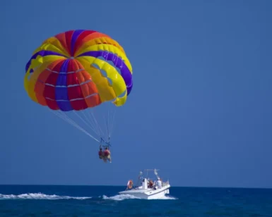 Una persona disfruta de la experiencia de Parasailing en Marbella, volando con un paracaídas vibrante, remolcado por un barco blanco sobre el océano brillante bajo un cielo azul claro. - ForTwo - Planes para parejas