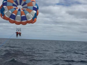 Dos personas disfrutan del Parasailing en Marbella, volando sobre las aguas azules con su vibrante paracaídas contrastando con el cielo nublado. - ForTwo - Planes para parejas