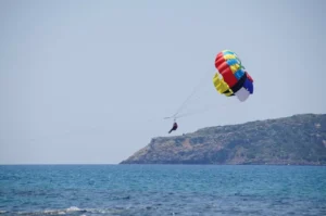 Una persona hace parasailing sobre el océano con un paracaídas multicolor cerca de una isla rocosa, experimentando Parasailing en Marbella. - ForTwo - Planes para parejas