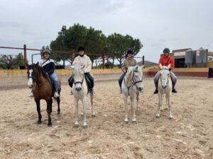 Cuatro jinetes, con casco y equipo, se sientan a lomos de sus caballos en el picadero al aire libre de Llíria, rodeados de frondosos árboles. Experimente el encantador "Paseo a caballo en Llíria: un plan romántico en la naturaleza" y disfrute de la belleza natural. - ForTwo - Planes para parejas