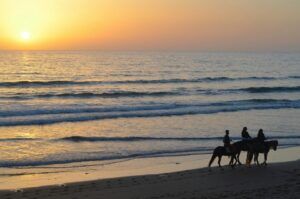 Tres personas disfrutan de un paseo a caballo al atardecer por la playa de Xeraco, con suaves olas de fondo, cortesía de Paseo a Caballo por la Playa de Xeraco. - ForTwo - Planes para parejas