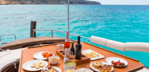 Una mesa en un barco con vino, aperitivos y pasteles con el impresionante telón de fondo del mar turquesa y la costa rocosa de la Costa de Palma durante el Paseo en Barco por la Costa de Palma. - ForTwo - Planes para parejas