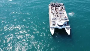 Vista aérea del "Paseo en catamarán por Valencia" con gente a bordo, navegando por las cristalinas aguas azules mientras los bañistas disfrutan de su tranquilo paseo cercano. - ForTwo - Planes para parejas