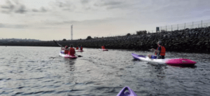Los kayakistas disfrutan de la tranquila experiencia del "Paseo en kayak por Getxo", navegando por aguas tranquilas cerca de Getxo y pasando por un terraplén rocoso bajo un cielo nublado. - ForTwo - Planes para parejas