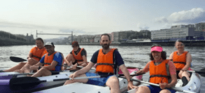 Seis personas, con chalecos salvavidas, navegan en kayak por un río de Getxo con el paisaje urbano de fondo durante el "Paseo en kayak por Getxo". - ForTwo - Planes para parejas