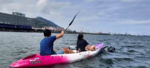 Dos personas en un kayak rosa disfrutan de la tranquilidad de las aguas de Getxo, dando un tranquilo "Paseo en kayak por Getxo" cerca de la ciudad costera bajo un cielo nublado. - ForTwo - Planes para parejas