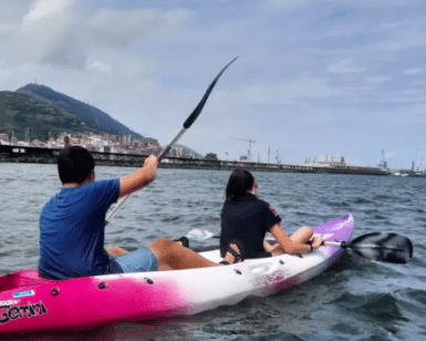 Dos personas en un kayak rosa disfrutan de la tranquilidad de las aguas de Getxo, dando un tranquilo "Paseo en kayak por Getxo" cerca de la ciudad costera bajo un cielo nublado. - ForTwo - Planes para parejas
